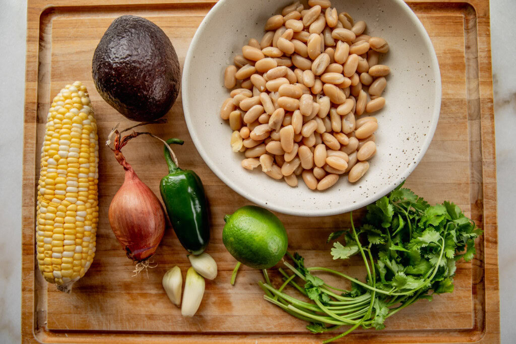 Cutting board with beans in a bowl, cilantro, lime, jalapeno, garlic, shallot, avocado, and corn.