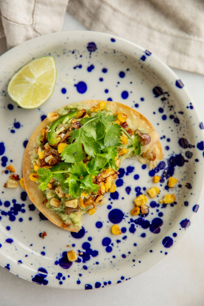 Half eaten tostada topped with mashed avocado and beans, corn, and cilantro on a speckled plate.