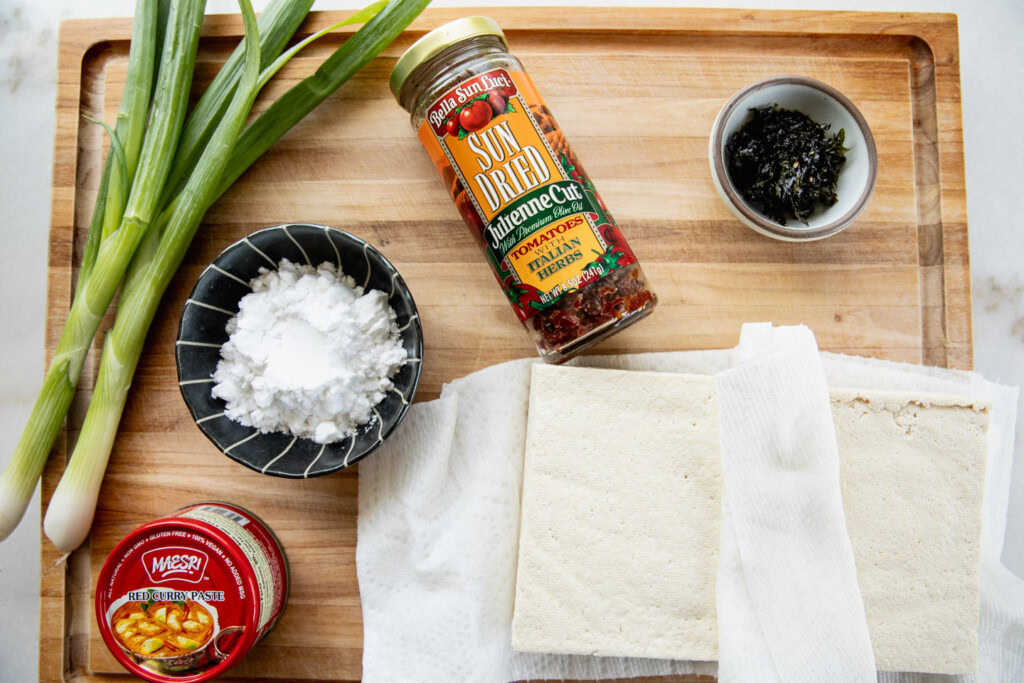 Cutting board topped with tofu, potato starch, thai curry, seaweed, scallions, and sun-dried tomatoes.