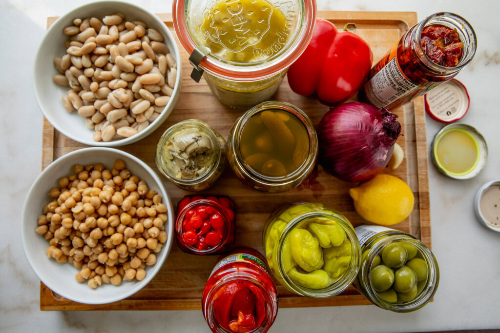 Bowls of beans, jared tofu feta, peppers, artichoke hearts, pepperoncini, sun-dried tomatoes, lemon, and olives on a cutting board.