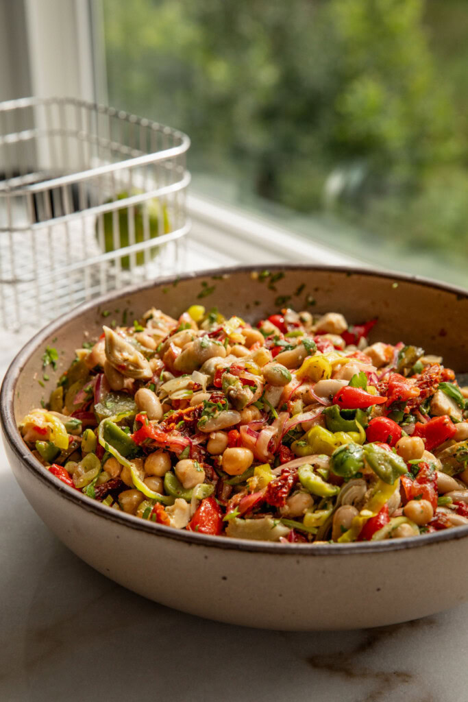 Mixing bowl filled with a tossed antipasto bean salad positioned by a window.