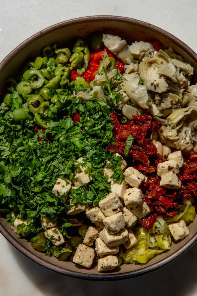 Sliced, olives, peppers, artichoke hearts, sun-dried tomatoes, feta, and herbs on top of beans in a bowl.
