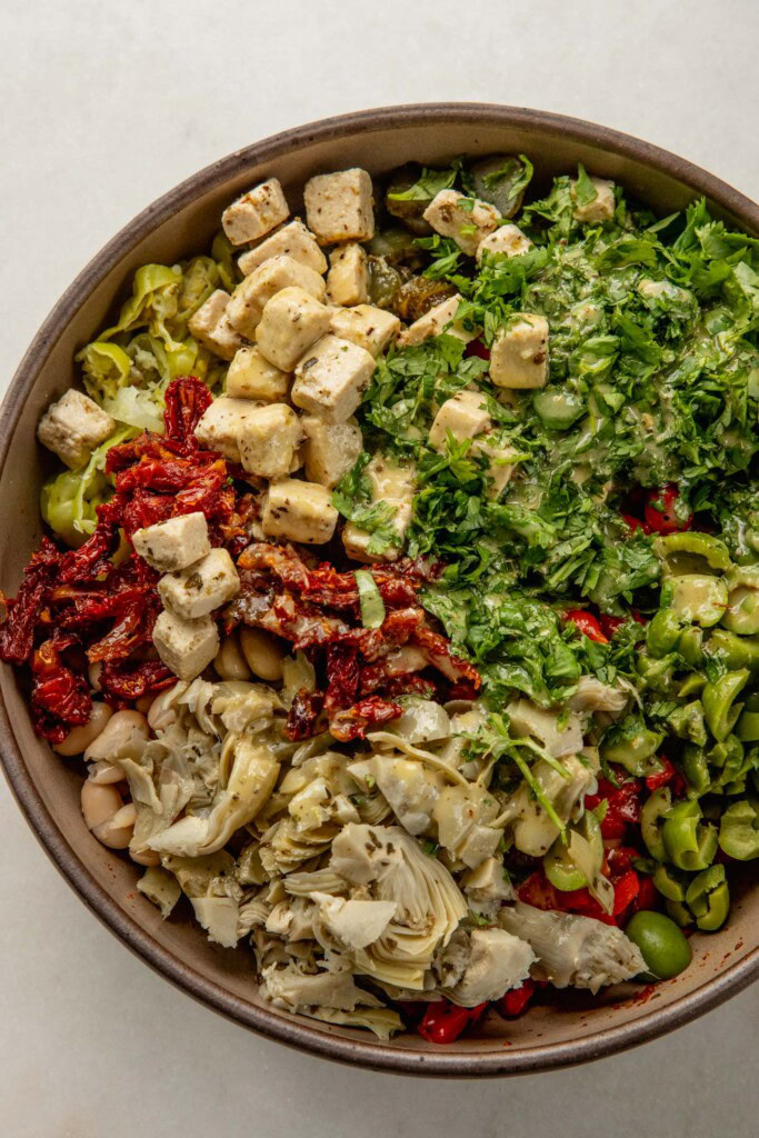 Sliced, olives, peppers, artichoke hearts, sun-dried tomatoes, feta, and herbs on top of beans in a bowl with the dressing poured on top.