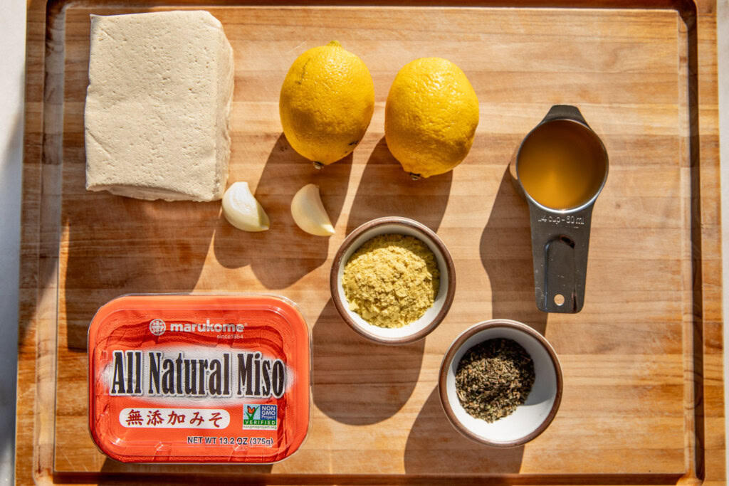 Cutting board with super firm tofu, lemons, garlic cloves, miso paste, nutritional yeast, apple cider vinegar, and dry herbs.