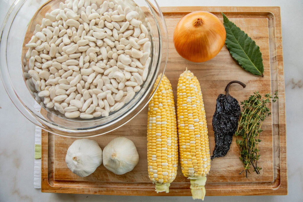 Cutting board topped with a bowl of soaked beans, ears of corn, dried ancho chili, bay leaf, thyme, onion, and garlic bulbs.