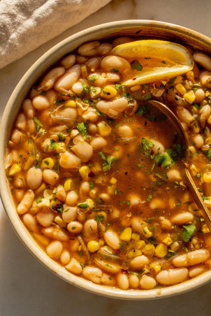 Moving the broth and beans around in a bowl with a gold spoon with a wedge of lemon tucked into the side.