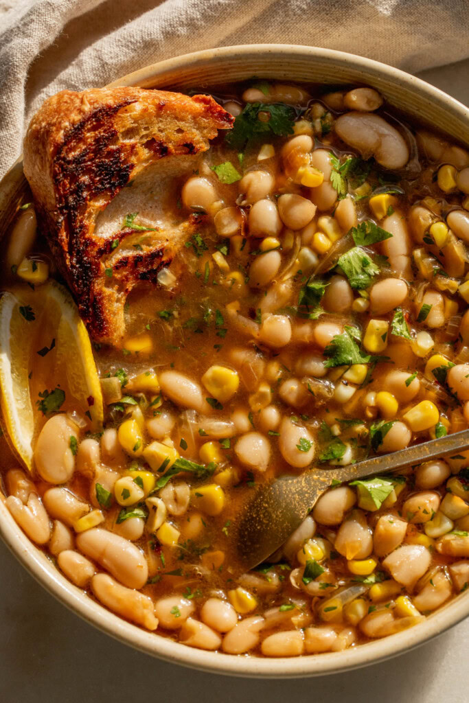 Bowl of brothy beans with a piece of bread tucked into the top left of the bowl.