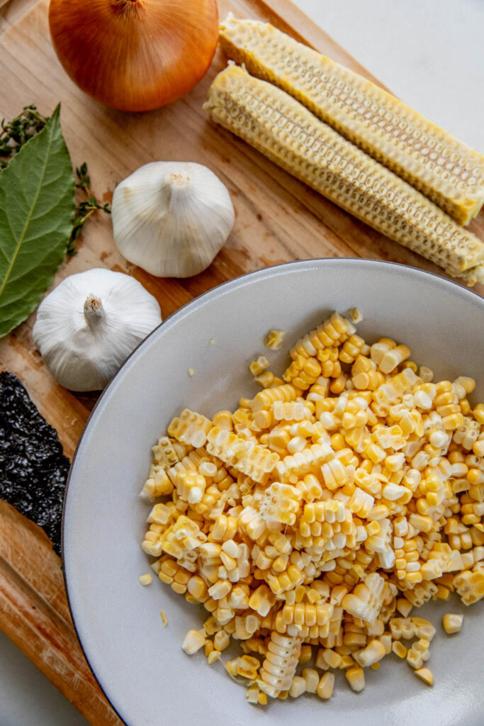 Cutting board topped with a bowl of corn kernels, corn cobs, garlic bulbs, bay leaf and dry chilies.