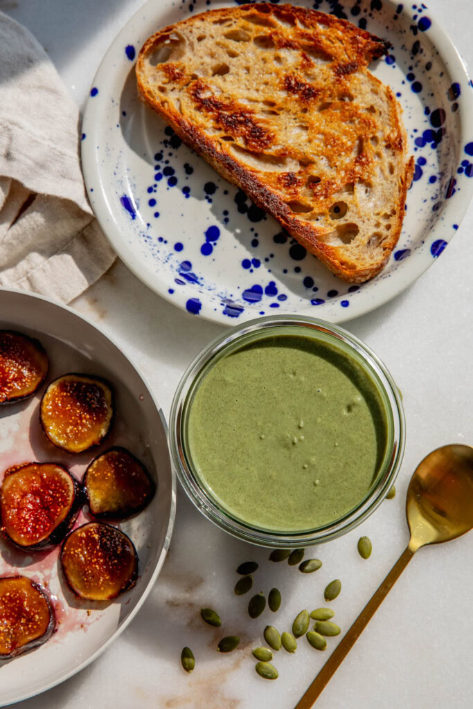 Jar of pumpkin seed butter next to a plate of toast and a pan filled with caramelized figs.