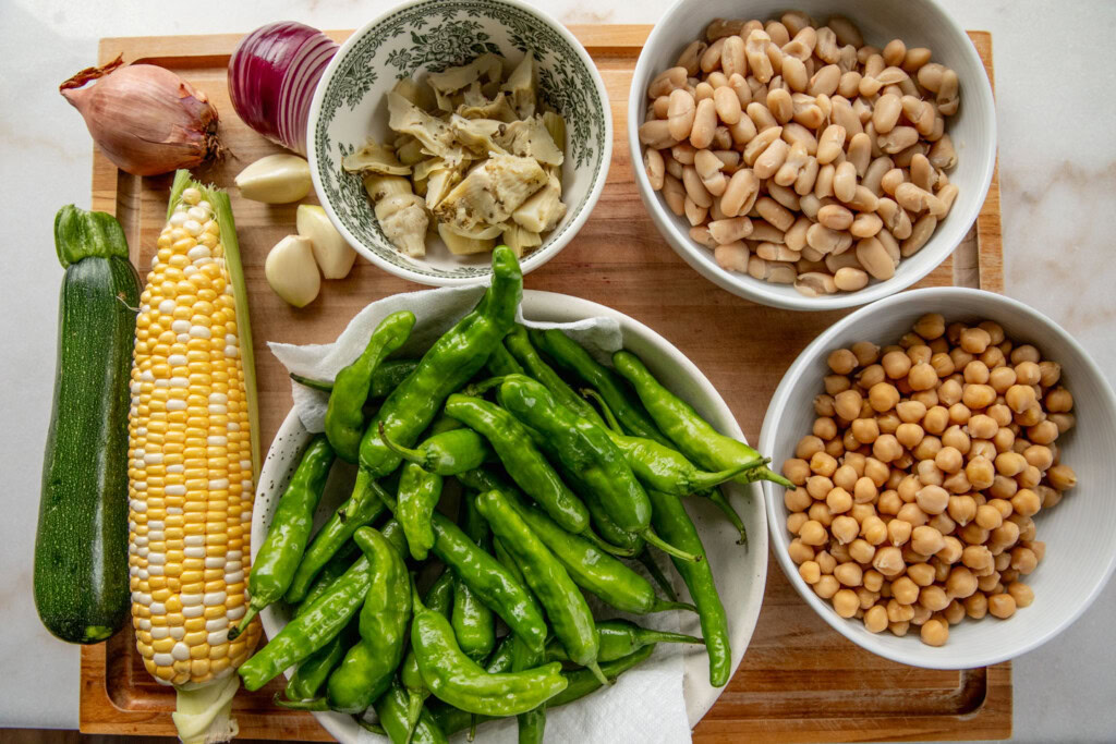 A cutting board topped with chickpeas, cannellini beans, shishito peppers, corn, shallots, garlic, and jalapeño.