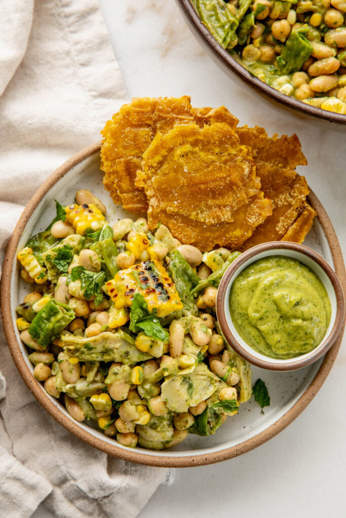 Plate filled with charred corn bean salad served with 3 baked plantains and a small bowl of extra salsa.