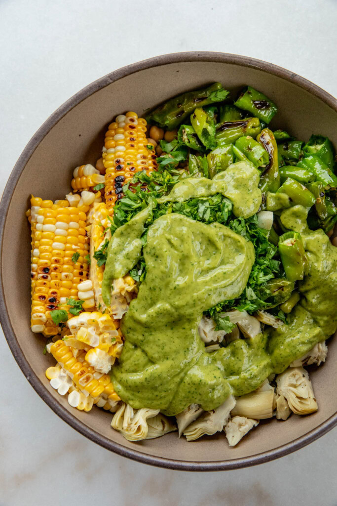 Large bowl of beans, corn, shishito peppers, artichoke hearts, cilantro with zucchini salsa poured overtop.