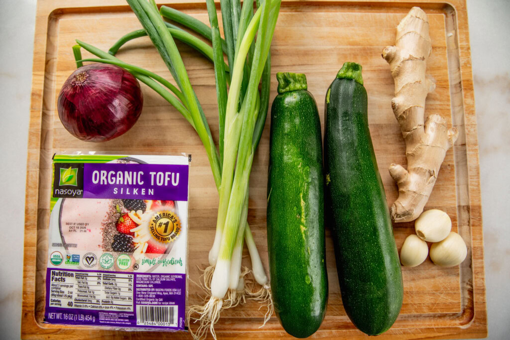 Cutting board with silken tofu, onion, zucchinis, garlic, scallions, and ginger.