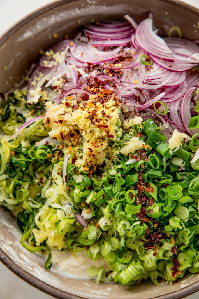 Adding shredded zucchini, onion, scallions, ginger, and garlic to a bowl with batter.