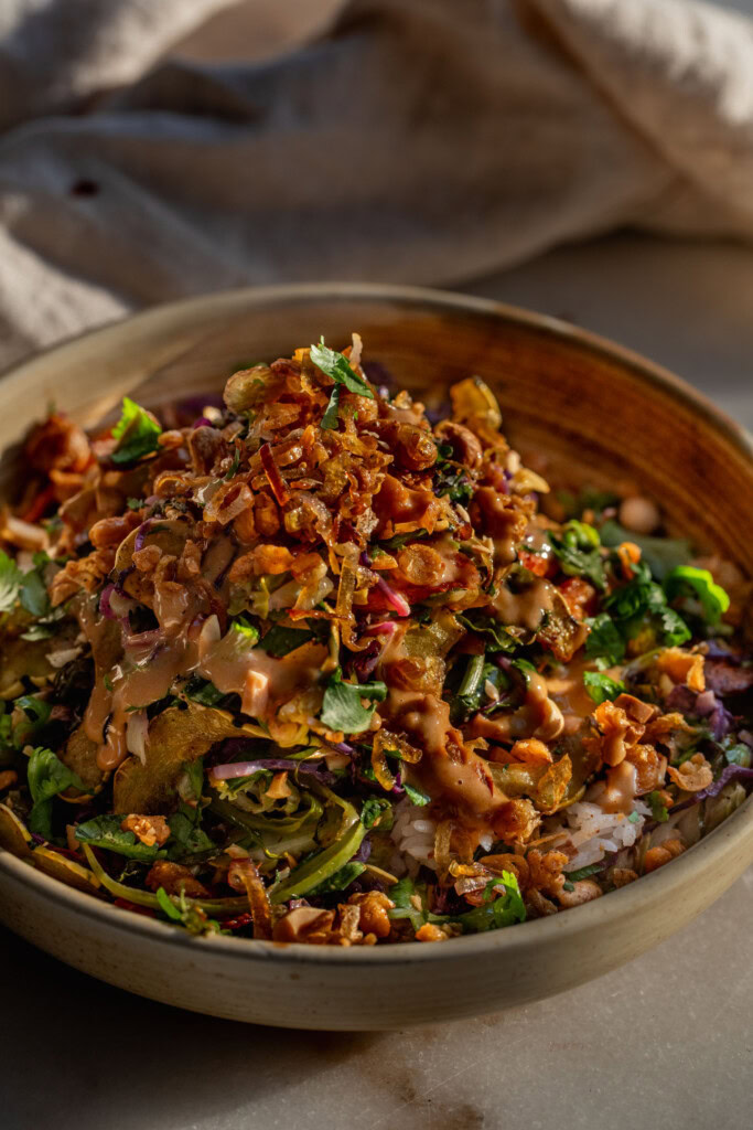 Close up of a bowl filled with rice, a baked mixed salad, chickpeas, shallots, and peanut sauce.