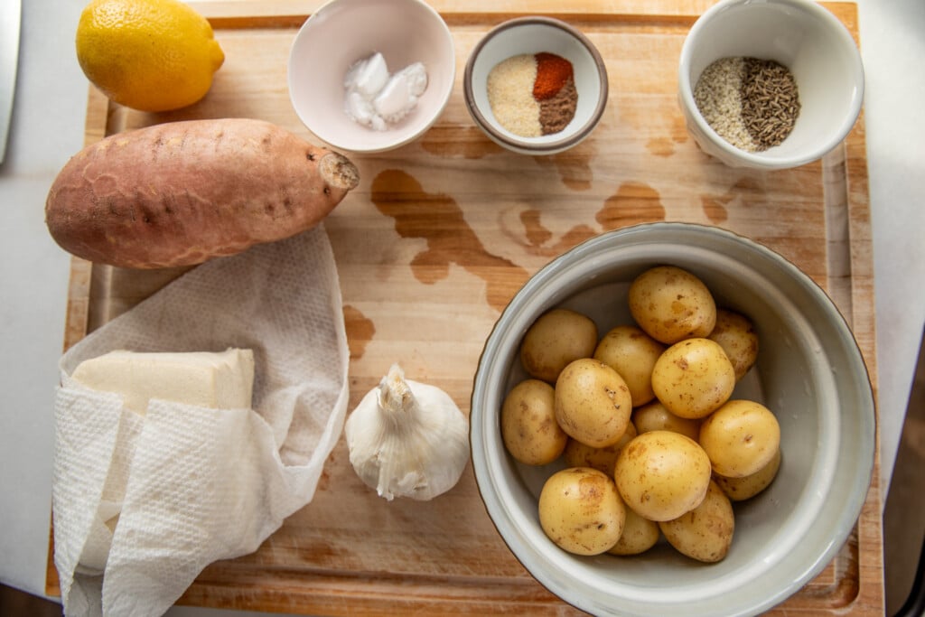Cutting board with bowl of baby potatoes, sweet potato, seasoning in bowls, lemon, potato starch, tofu, and garlic.