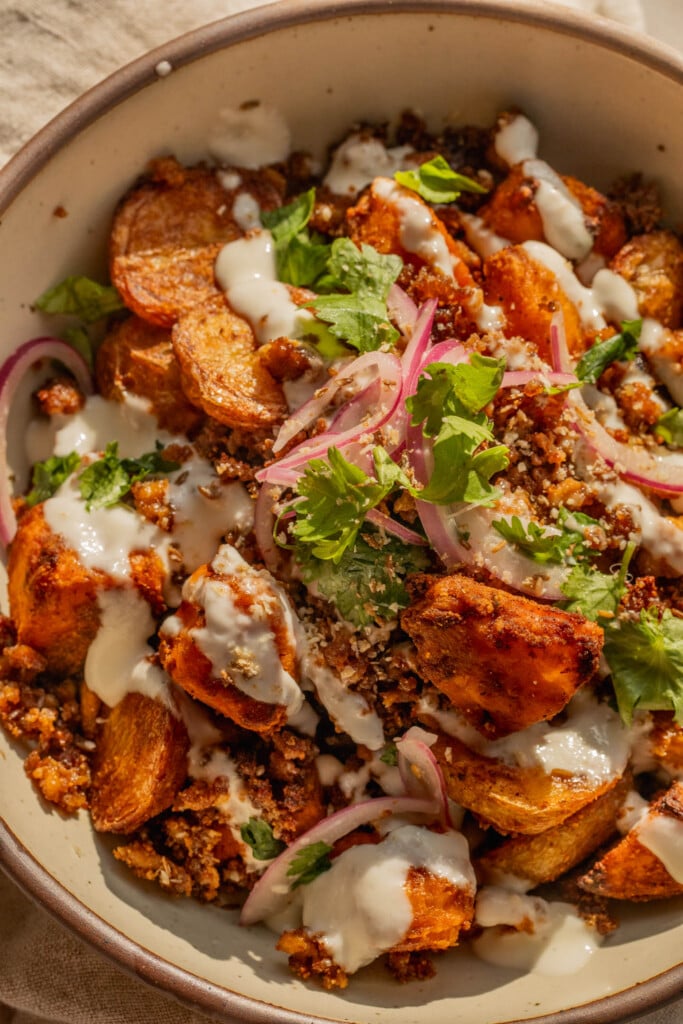 Close up of a bowl filled with roasted potatoes, crumbled tofu, garlic sauce, and marinated onions.